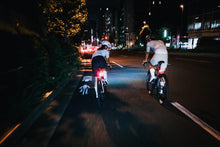 Load image into Gallery viewer, Two cyclists ride away on a city street at night, viewed from behind. Their rear bike lights glow red and white, illuminating the road, visible in the background.